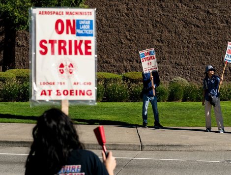 A WOMAN HOLDING A SIGN SAYING ON STRIKE AT BOEING AND A RED COWBELL FACES OTHER STRIKE WORKERS ACROSS A STREET HOLDING SIMILAR SIGNS